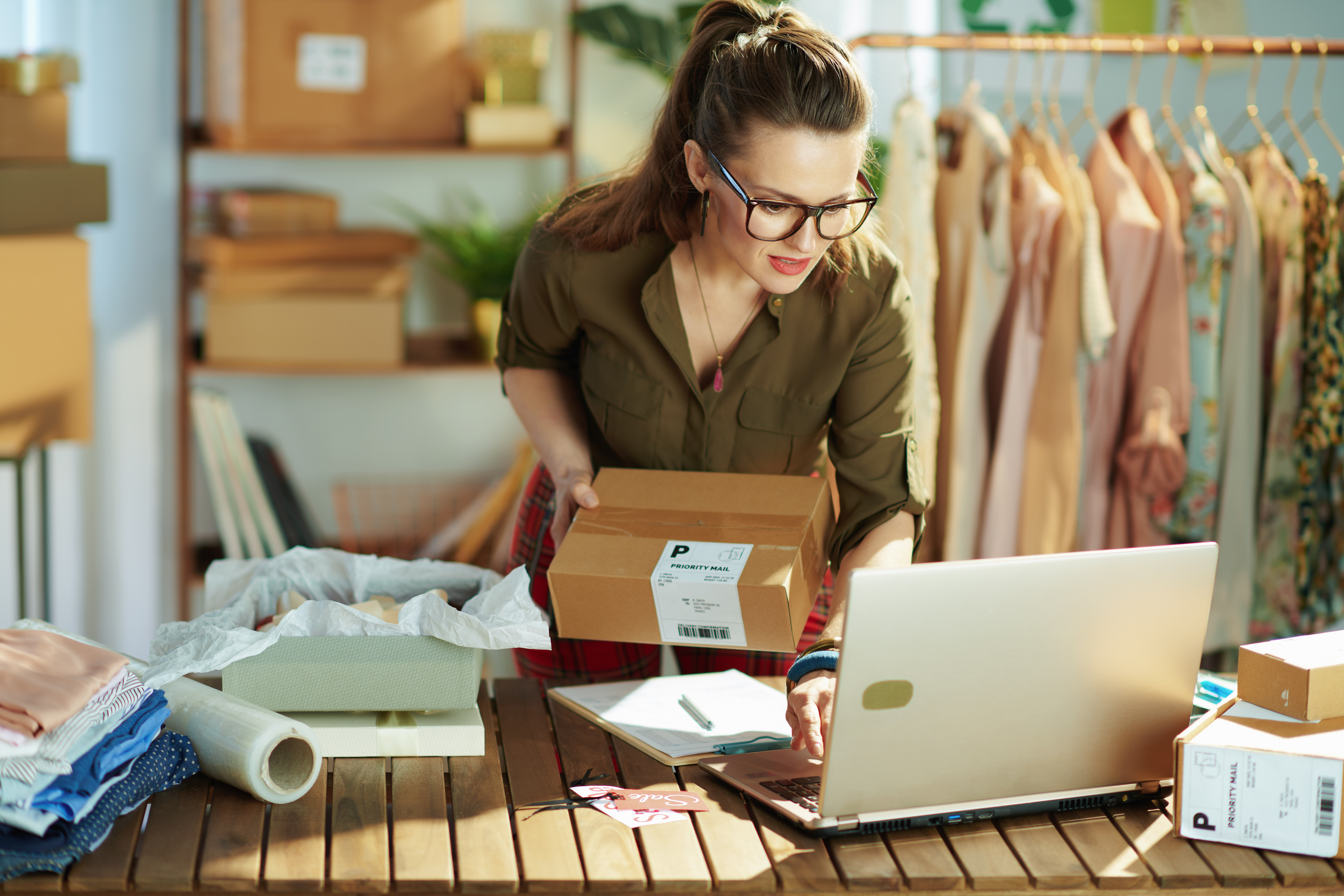 Eine Frau mit Brille steht vor einem Laptop und hat ein Paket in der Hand.