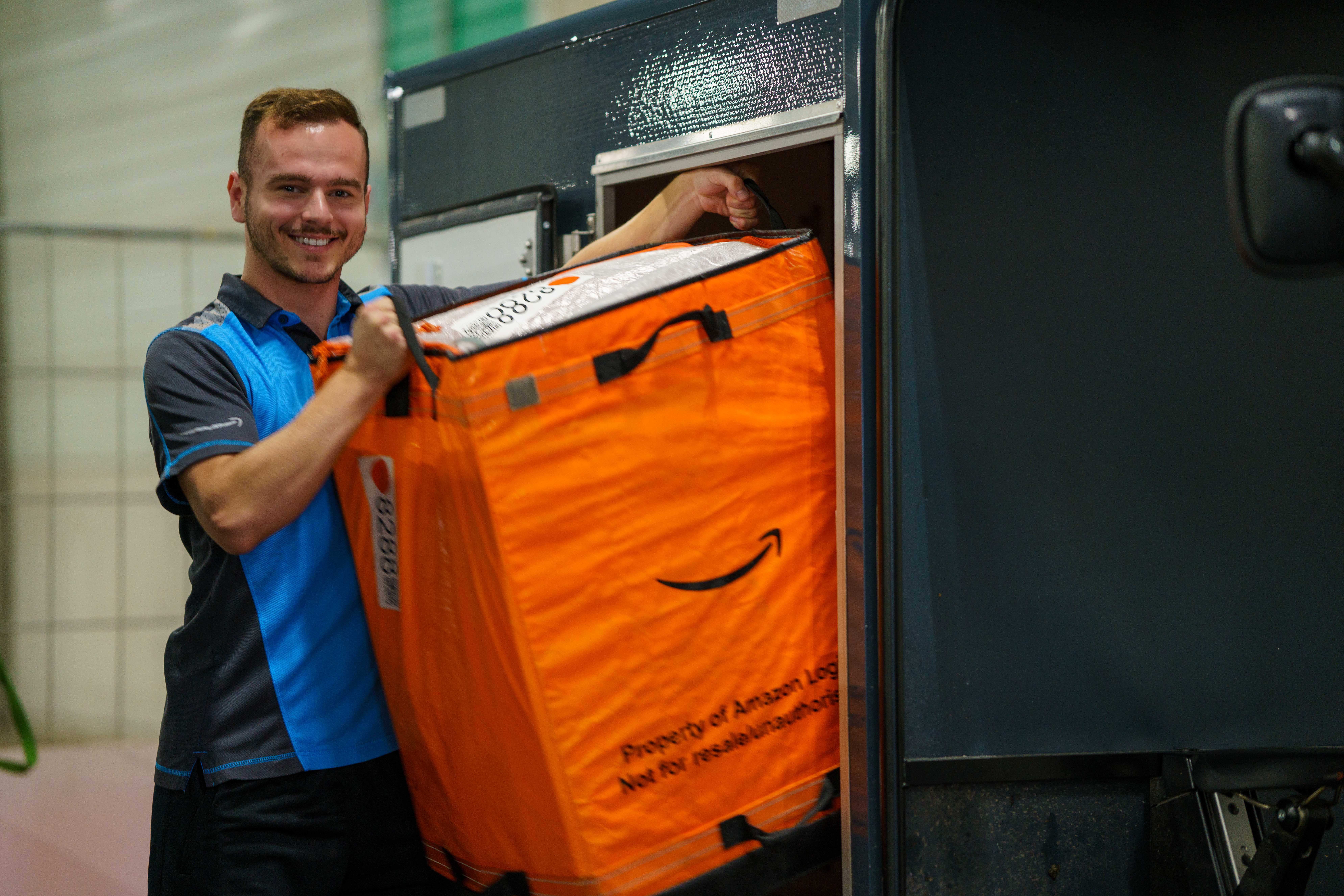 Ein junger Fahrer in blauer Dienstkleidung mit Amazon-Logo lädt eine orangene Versandtasche in ein E-Cargo Bike.