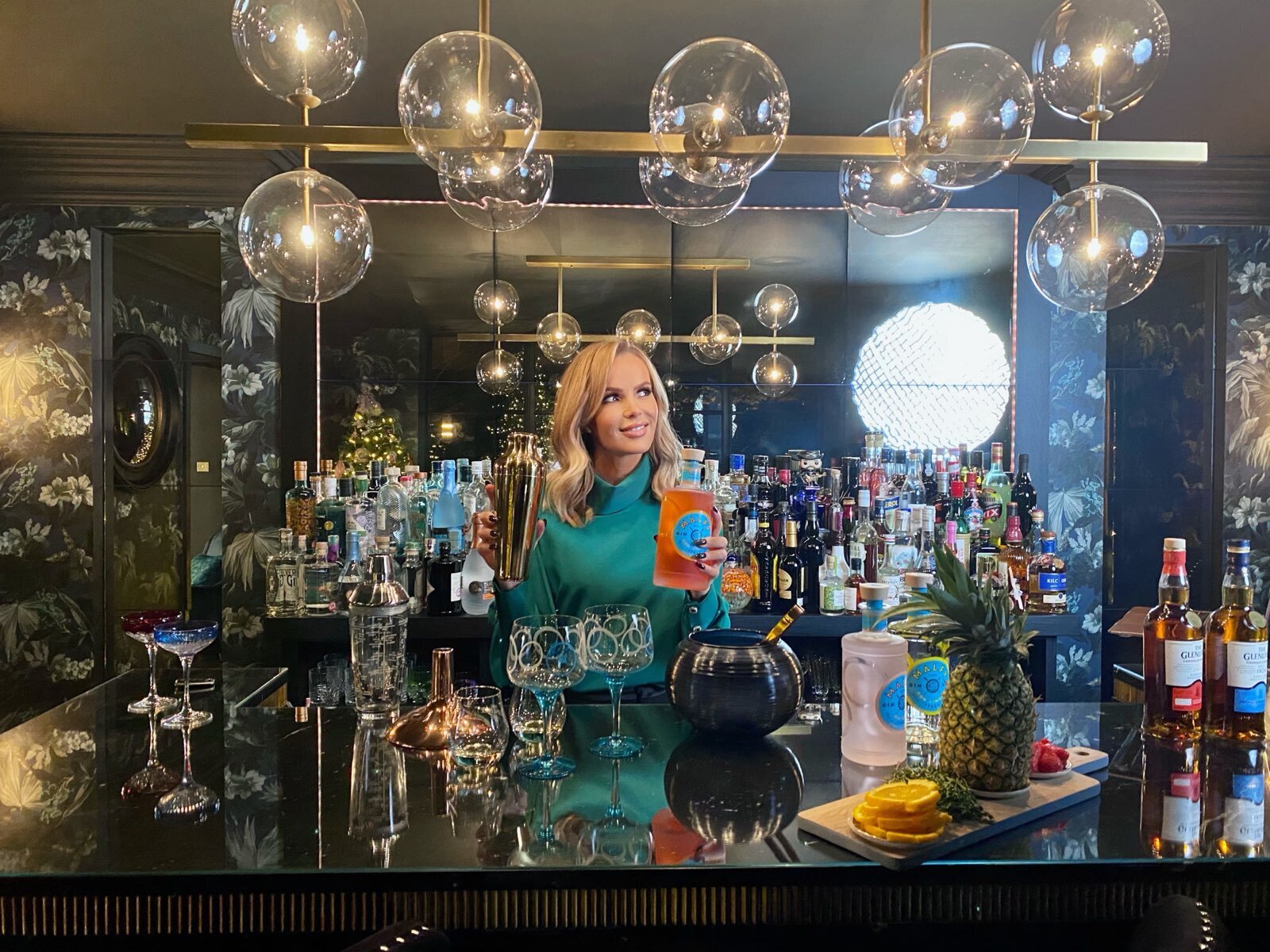 Amanda Holden holdng cocktail shaker sat at a bar surrounded by glass lighting