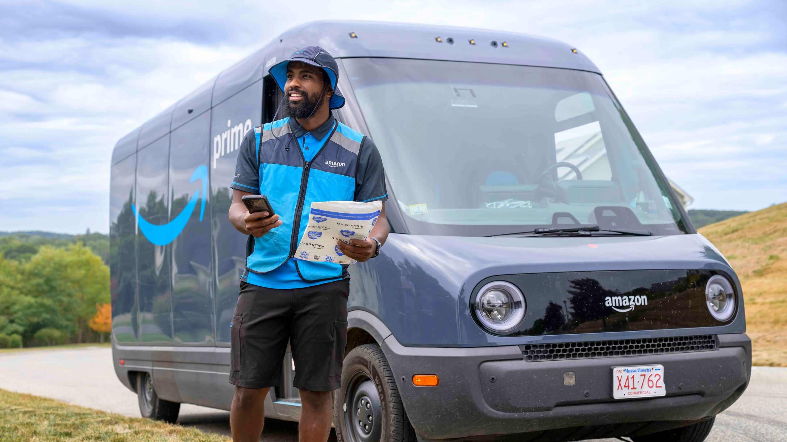 An Amazon delivery driver standing on the side of a road, holding a package in one hand and a mobile device in the other. Behind them is an Amazon delivery truck. 