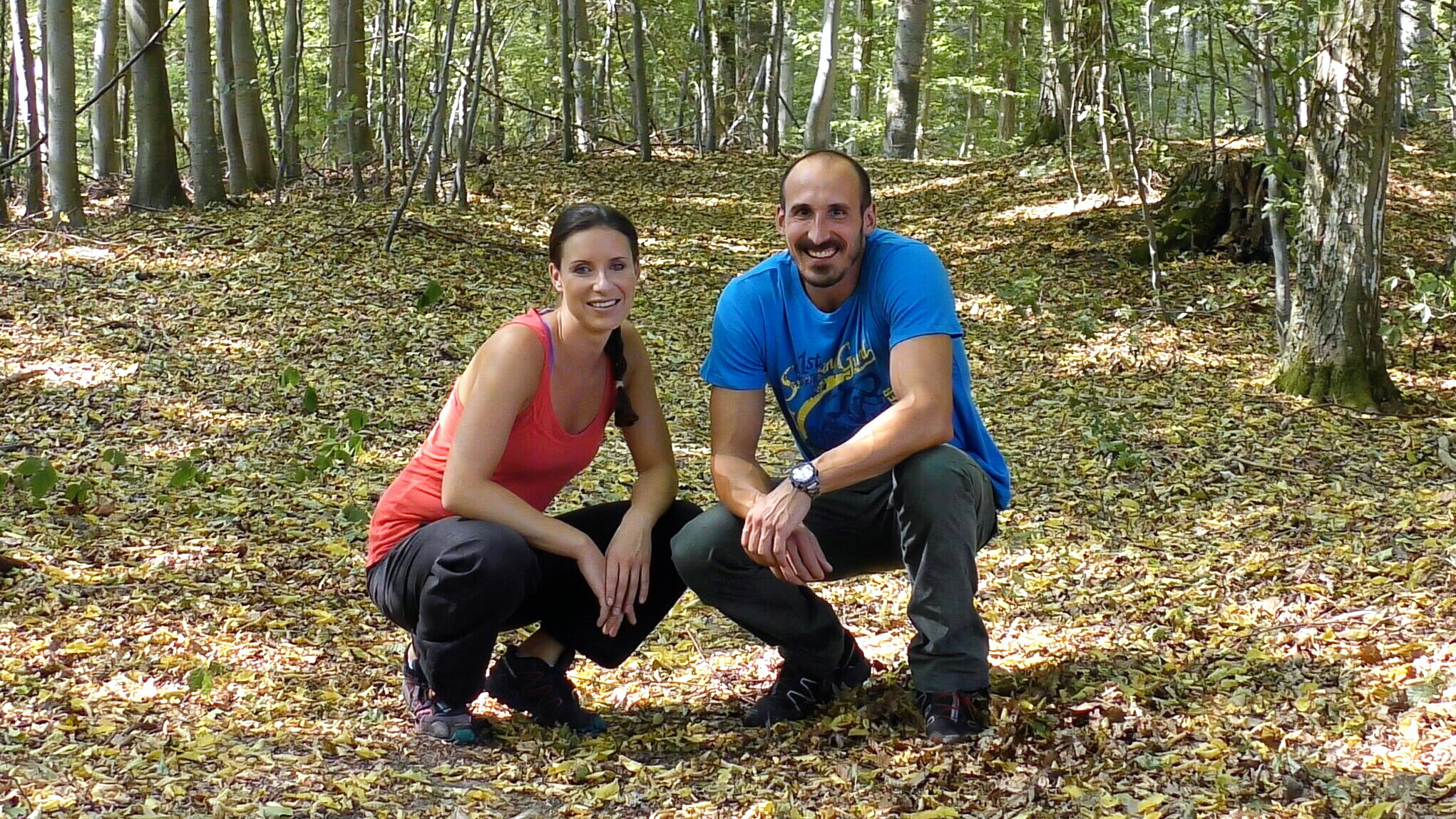 Ein Mann und eine Frau sitzen auf einem Waldboden und lachen in die Kamera.