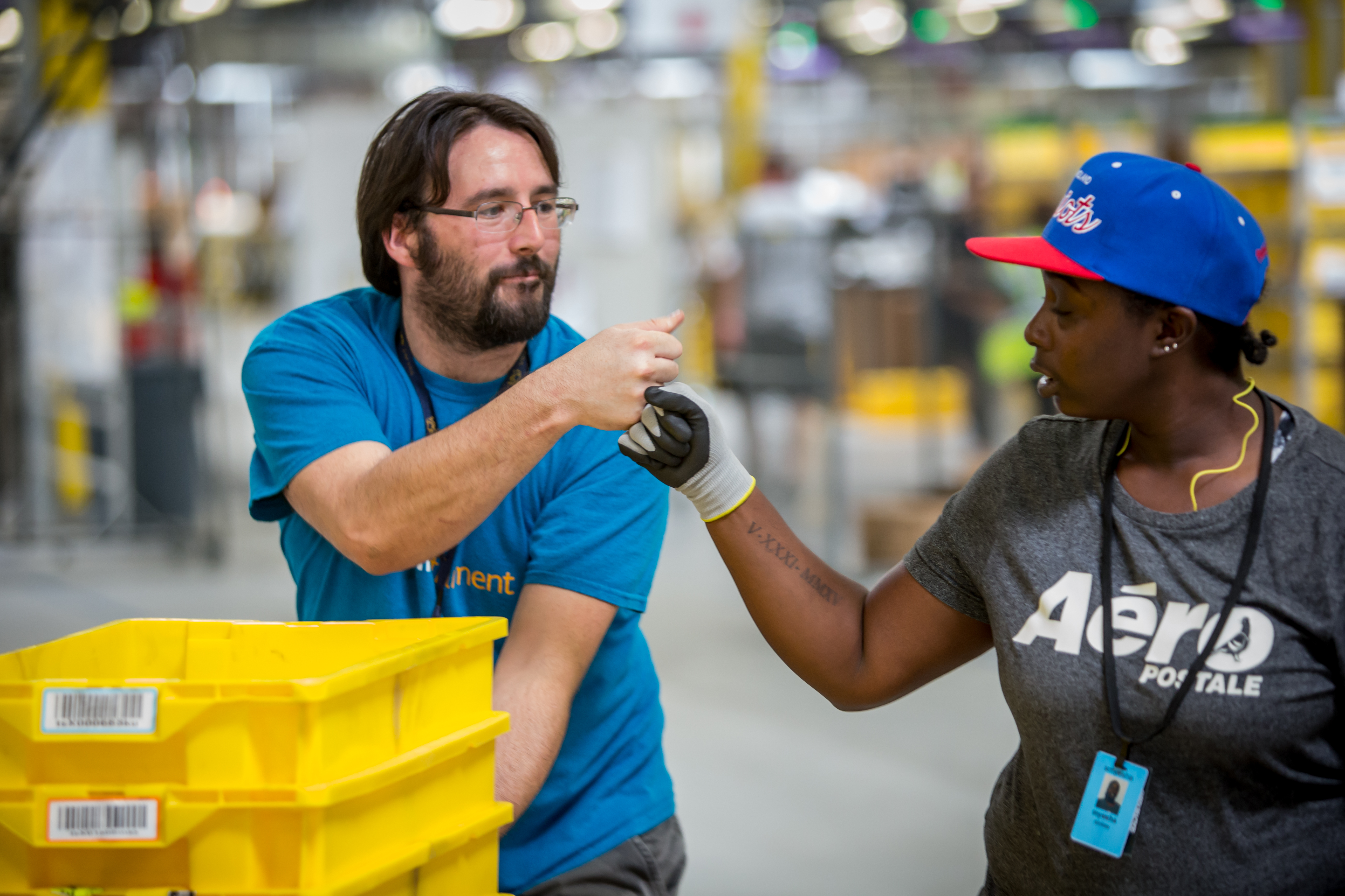 An male Amazon associate in a blue t-shirt bumps fists with a female Amazon associate wearing a grey t-shirt that says 'Aeropostale'. Three yellow bins are in front of the man, and the woman is wearing a blue baseball cap. 