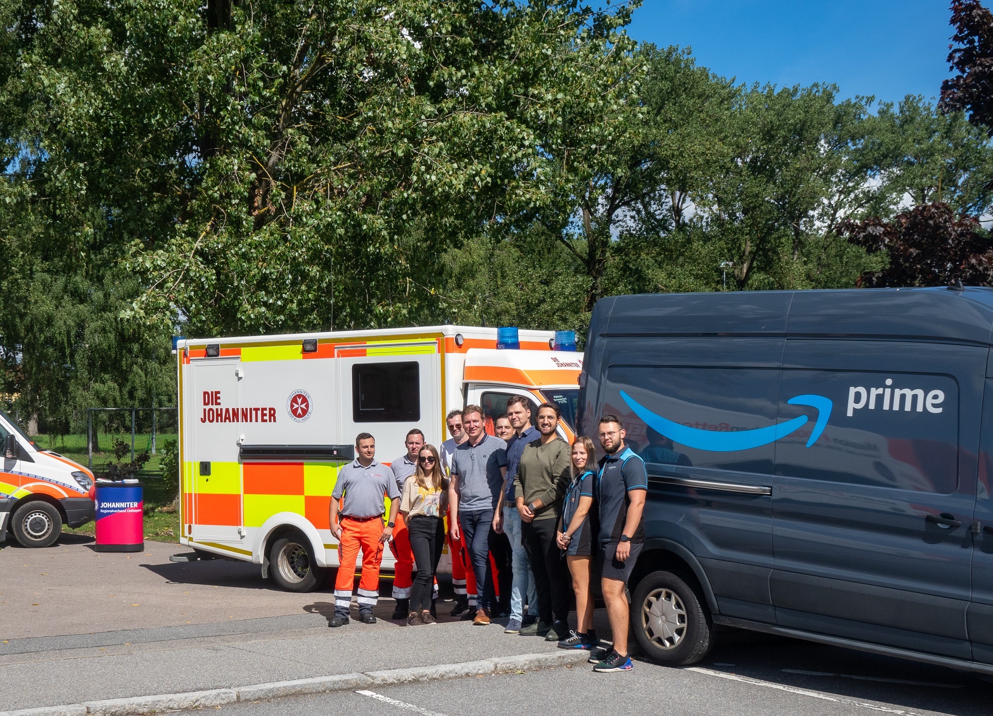 Amazon Lieferfahrerinnen und Lieferfahrer vor einem Amazon Prime Lieferfahrzeug zusammen mit Mitarbeitenden der Johanniter-Unfall-Hilfe vor einem Rettungwagen.