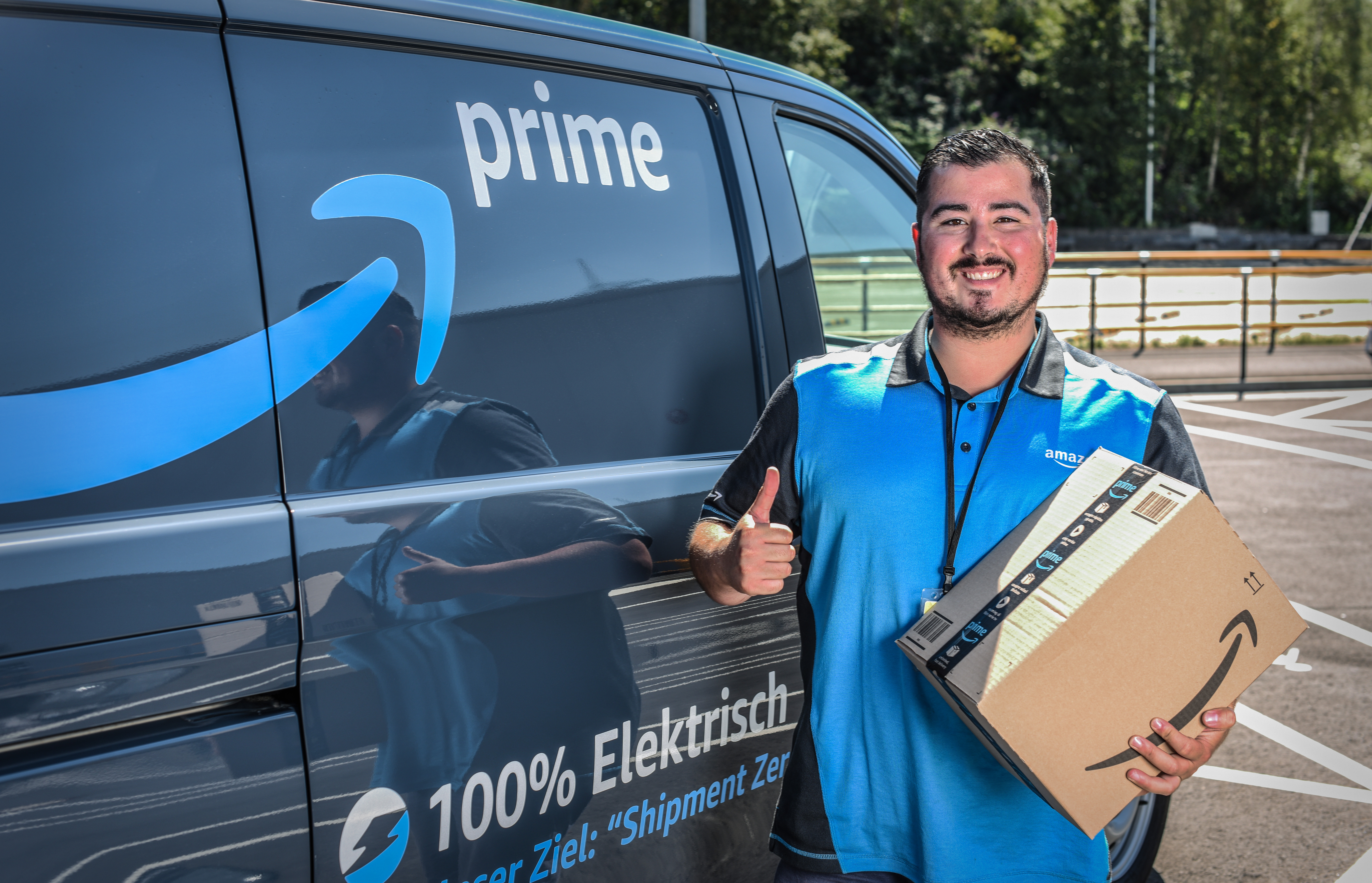 Ein lächelnder junger Mann in Amazon Shirt steht vor einem Prime Lieferfahrzeug. Er hält ein Amazon Paket in der Hand.