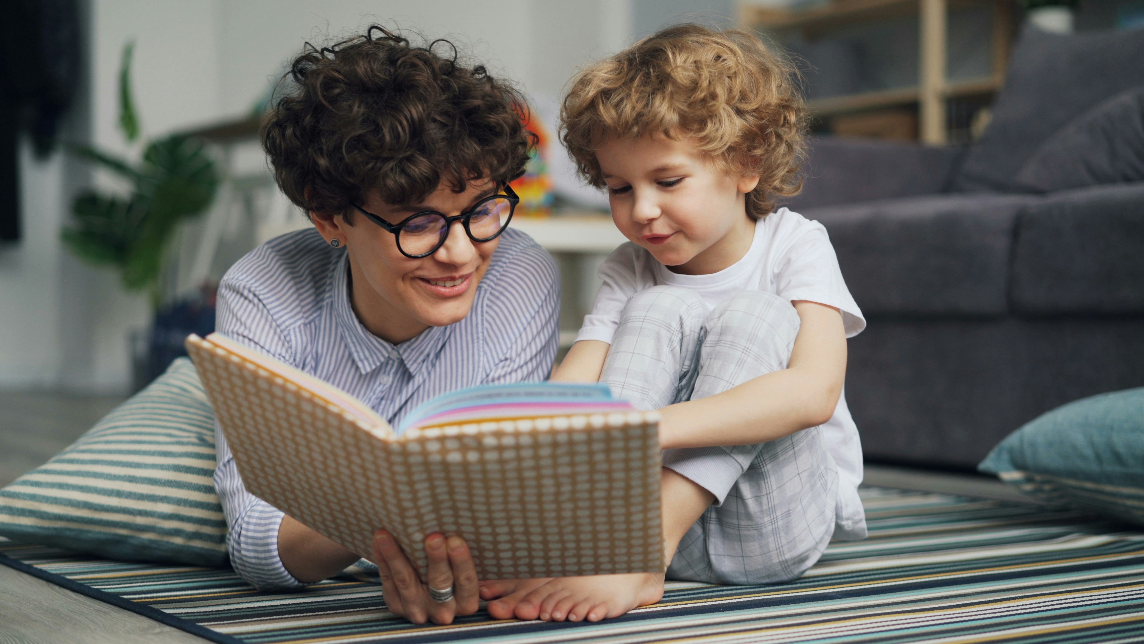 A parent with curly brown hair and round glasses reads a book to their child