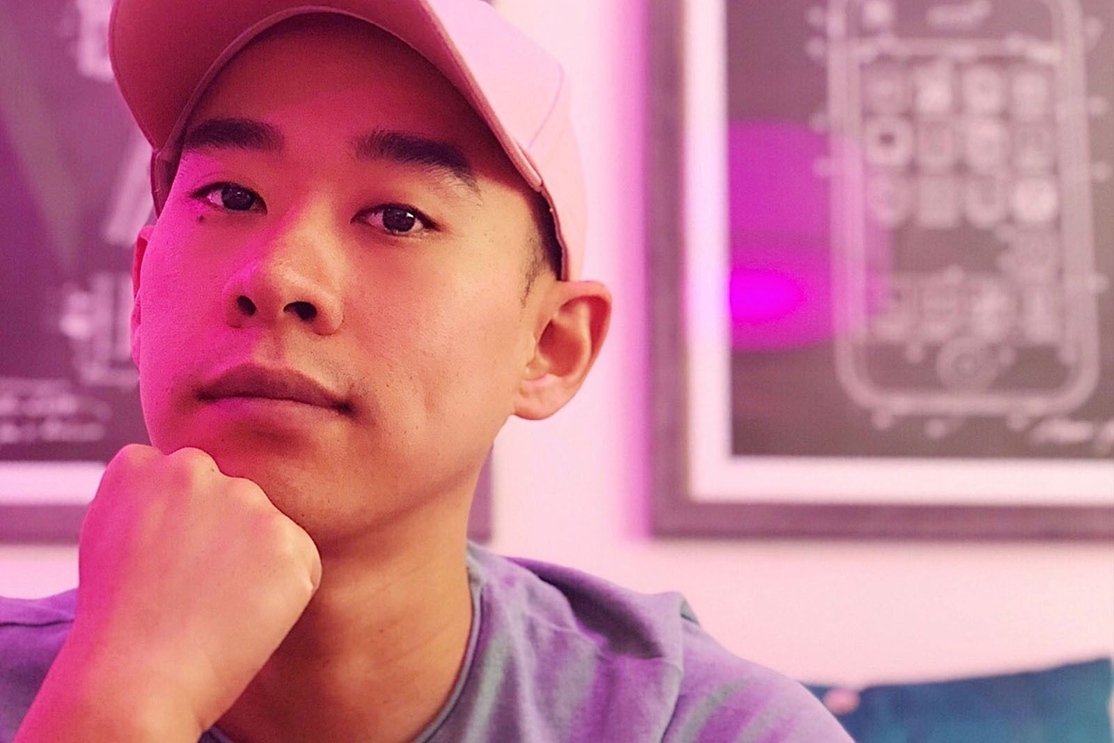 A headshot image of a man smiling softly for a photo under pink light. He has a white baseball hat on and has his chin rested on his fist.