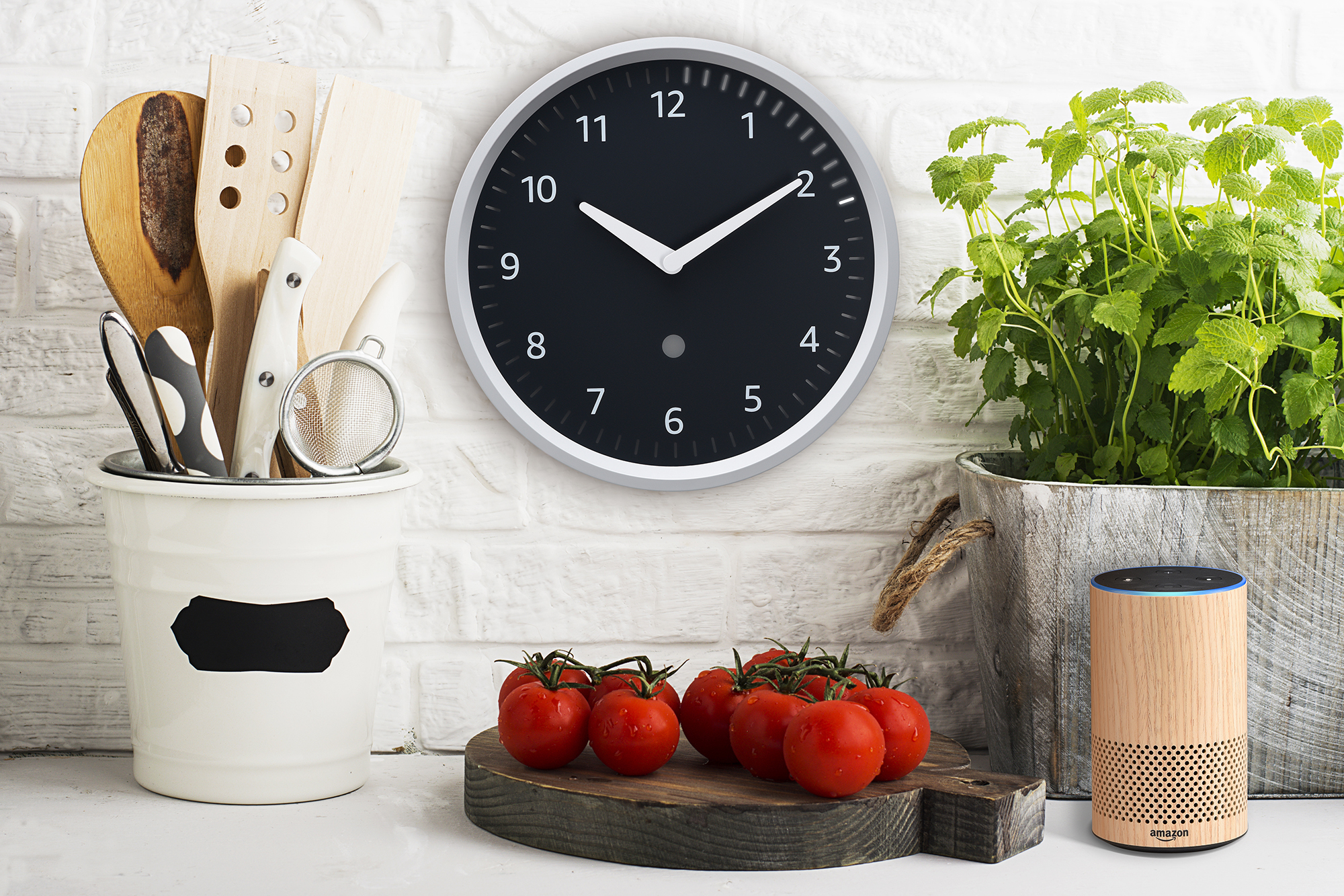 Amazon Echo Clock on the wall in a kitchen. To the left is a carafe of kitchen tools, below the clock is a cutting board with tomatoes. To the right, an herb growing in a container, and an Echo device. 