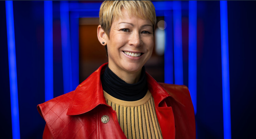 An image of a woman smiling for a headshot in a hallway of blue lights. She is wearing a red jacket and yellow sweater. 