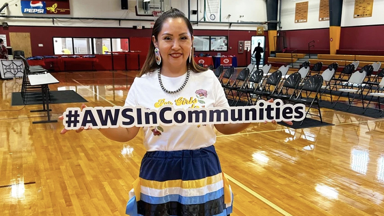 A photo of a member of the Confederated Tribes of Umatilla Indian Reservation standing in a gym, holding a sign that states, #AWSInCommunities.