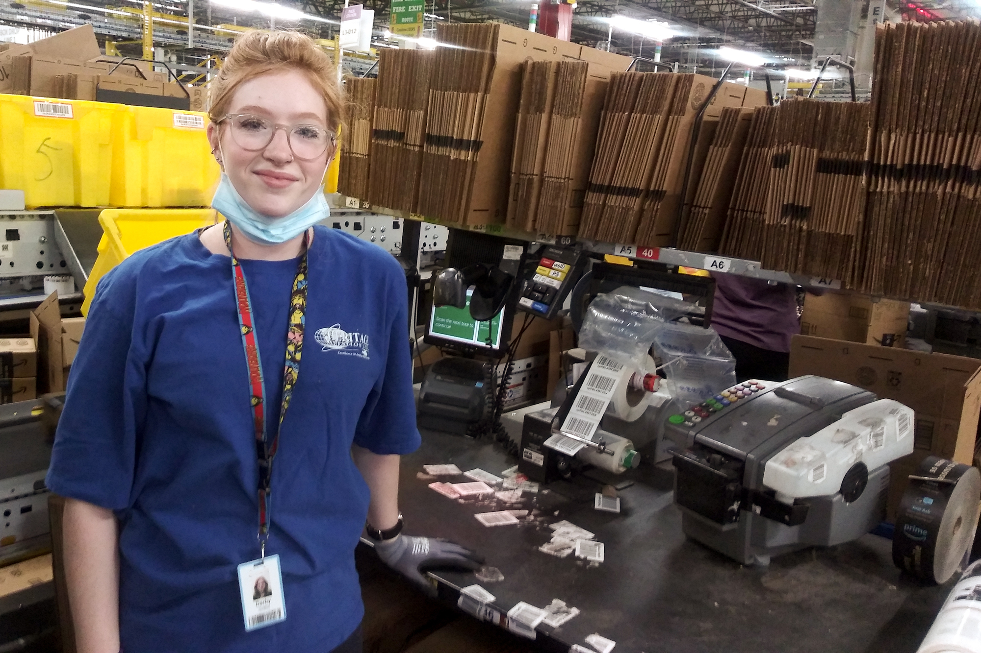 A woman stands at a workstation stocked with cardboard boxes and labeling machines. She has a protective mask on, ready to cover her mouth and nose.