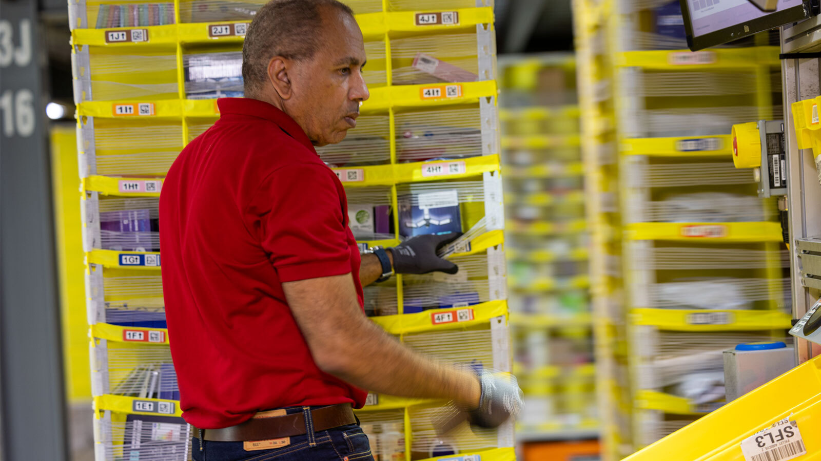 an image of an amazon employee placing items in a large yellow tote with multiple cubbies for customer orders. There are several totes in the background, being moved by small, orange robots underneath them.