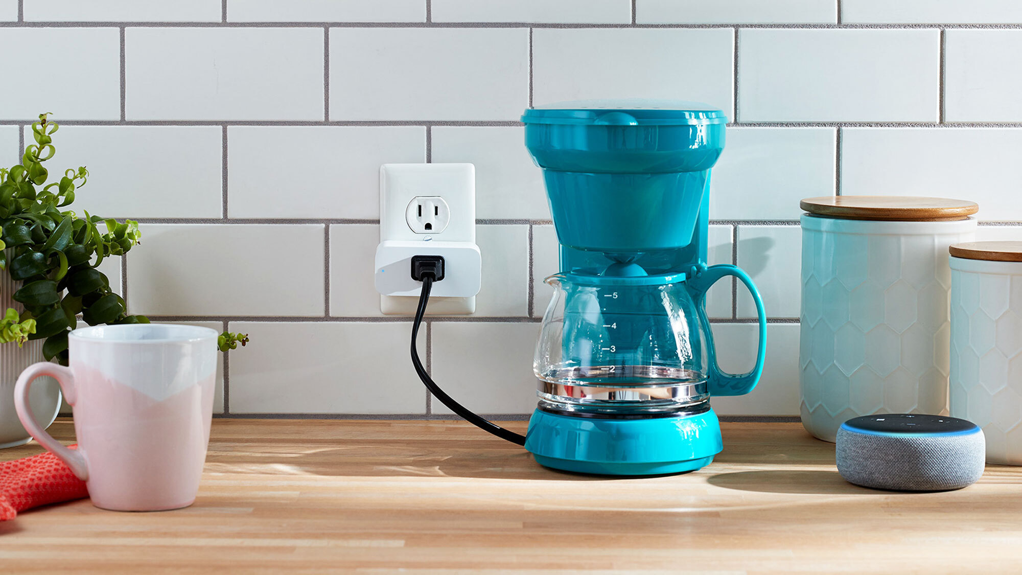 An image of a coffee maker plugged to a smart plug. There is an Echo device next to it and a white mug on the other side, along with miscellaneous items on the counter. 