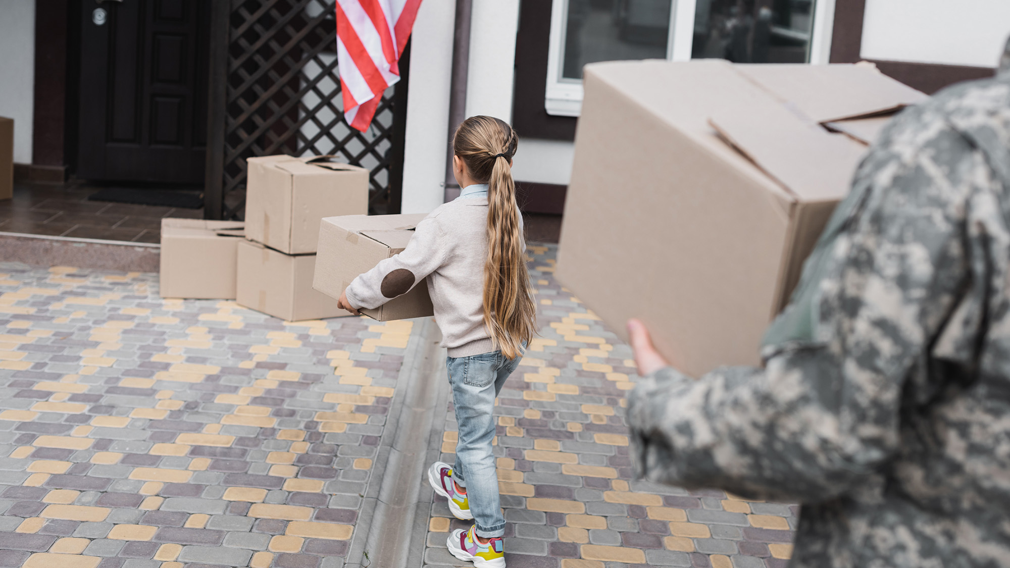 A child carries a moving box, behind her a man in military fatigues also carries a box. 