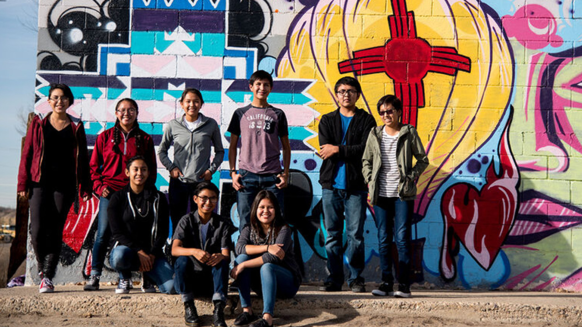 An image of a group of students standing and sitting in front of a graffiti wall. 