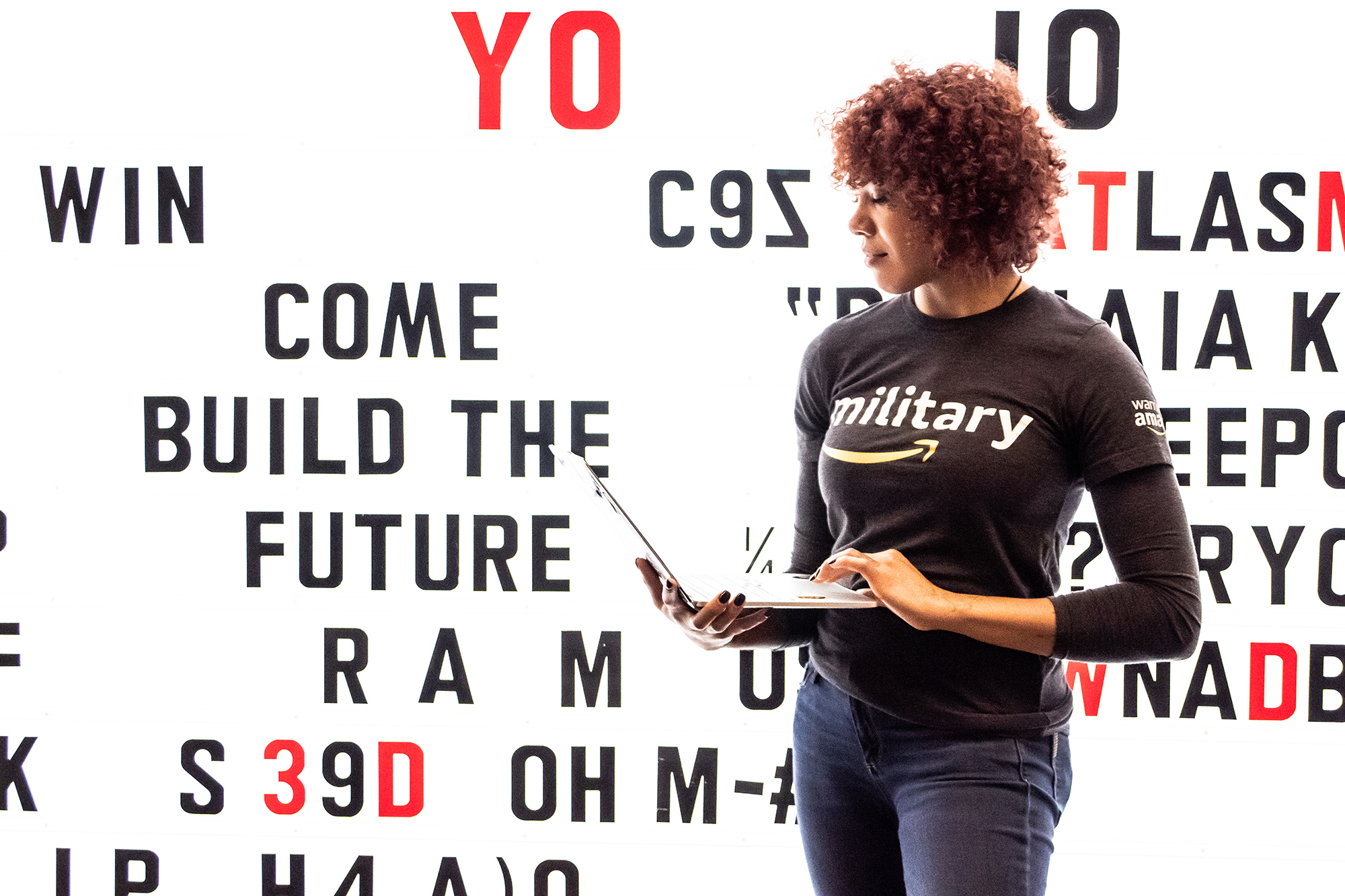 A woman stands in front of a lit wall, holding a laptop computer. 