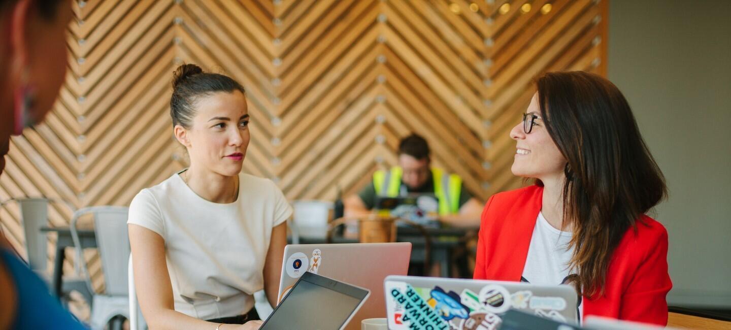 Two women work on their laptops and talk in an office setting.