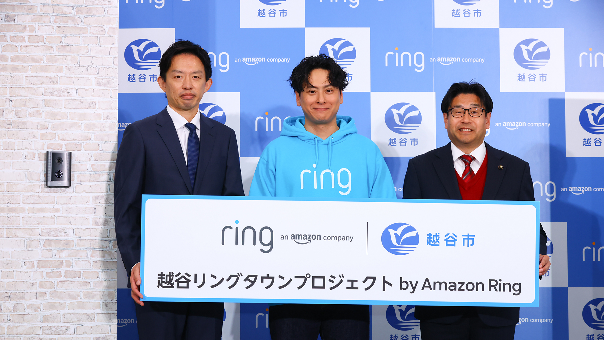 越谷市のロゴとringのロゴが市松模様になったパネルの前で、ボードを持って立つ3人の男性。ボードには「越谷リングタウンプロジェクト」の文字