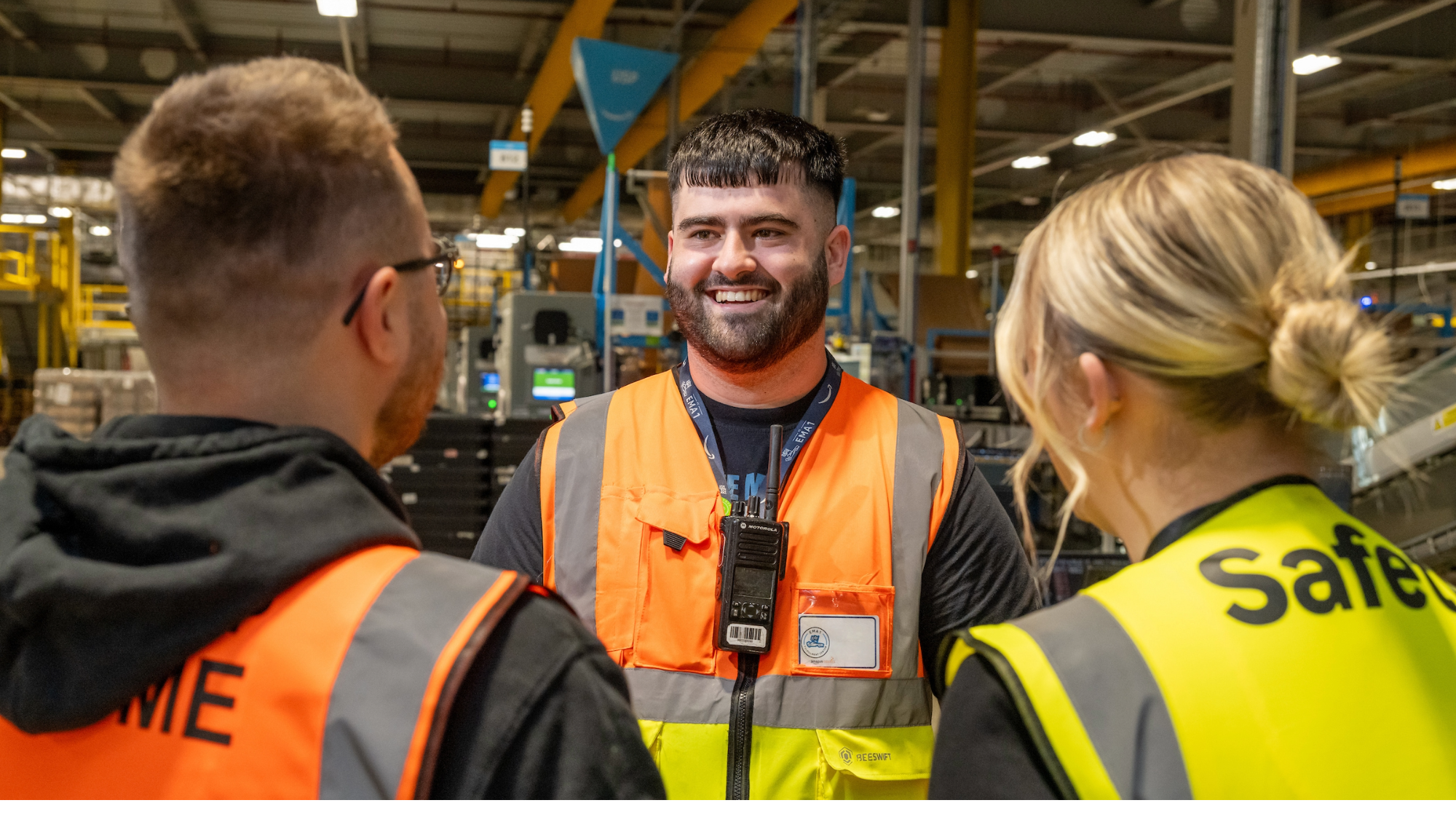 Fulfilment Center manager in the UK stands opposite two employees with a smile on this face. All three are wearing high-vis jackets