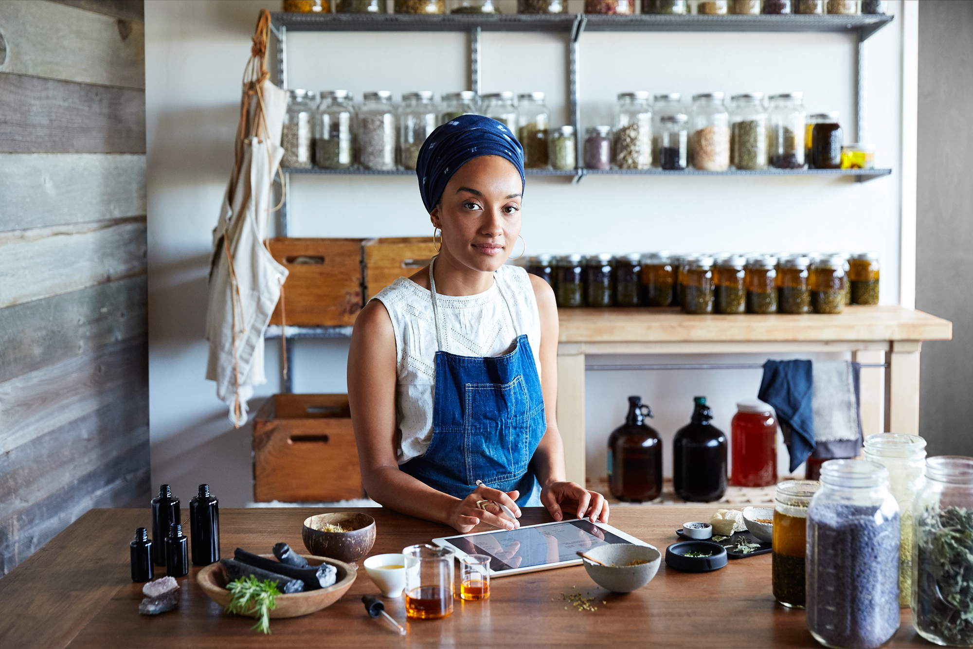 A woman stands at a table, working on an iPad. Behind her are jars of preserves or other homemade products, and there are herbs and ingredients in front of her. 