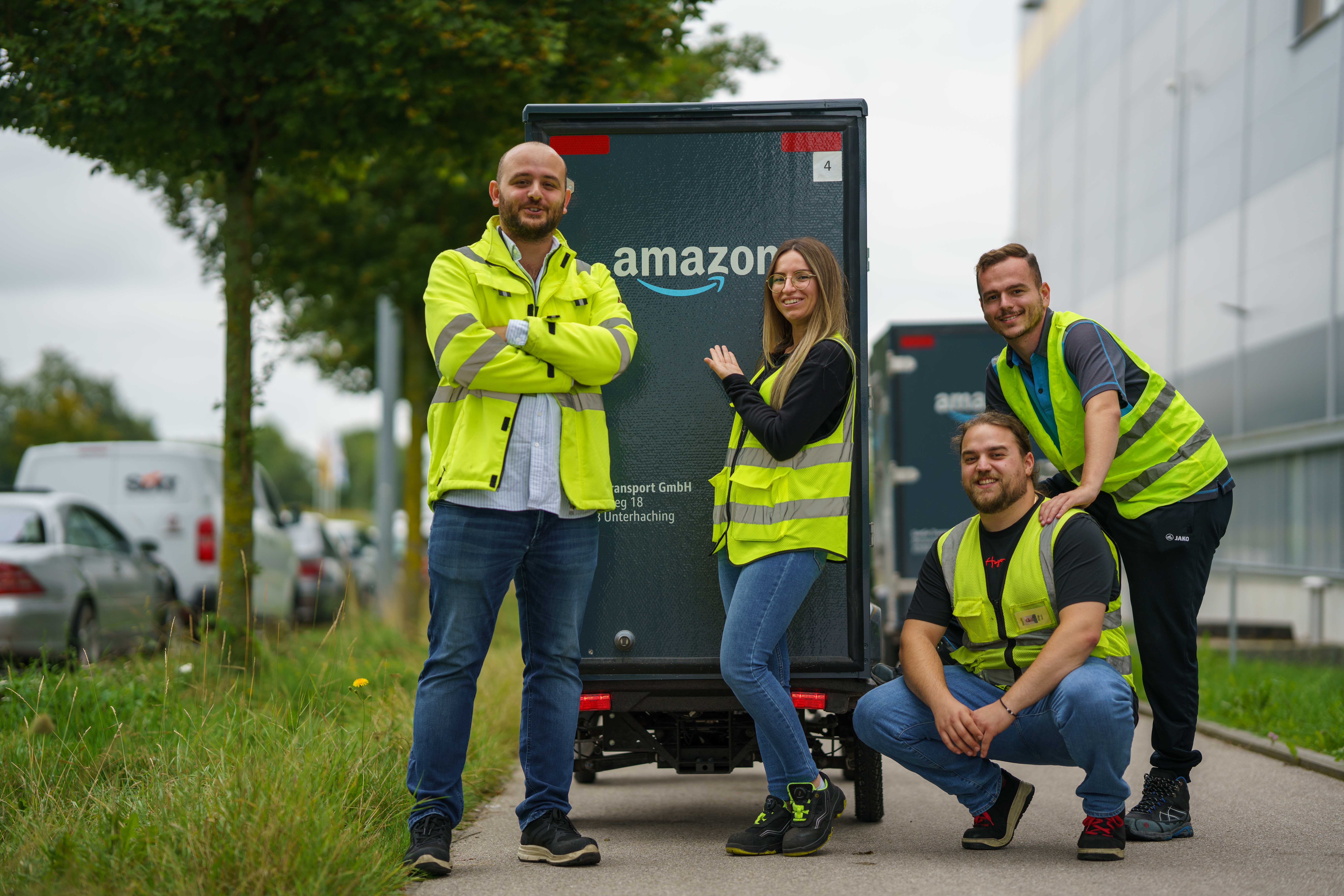 Gruppenbild im Grünen vor zwei E-Cargo-Bikes, auf denen man von hinten große Amazon-Logos erkennen kann: Alle Mitarbeiter tragen gelbe Sicherheitswesten und lächeln in die Kamera.