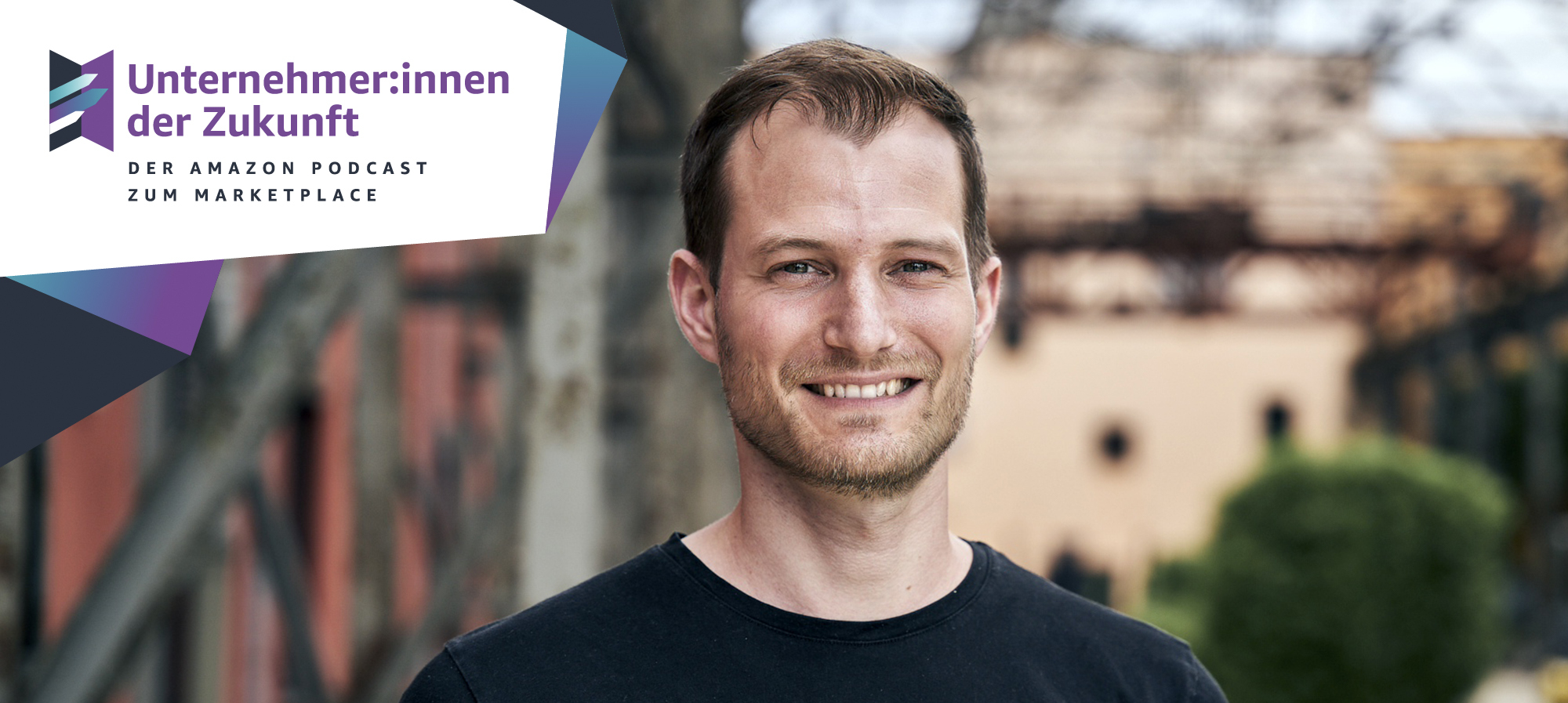 Ein Mann mit braunen Haaren und einem schwarzen T-shirt lacht in die Kamera