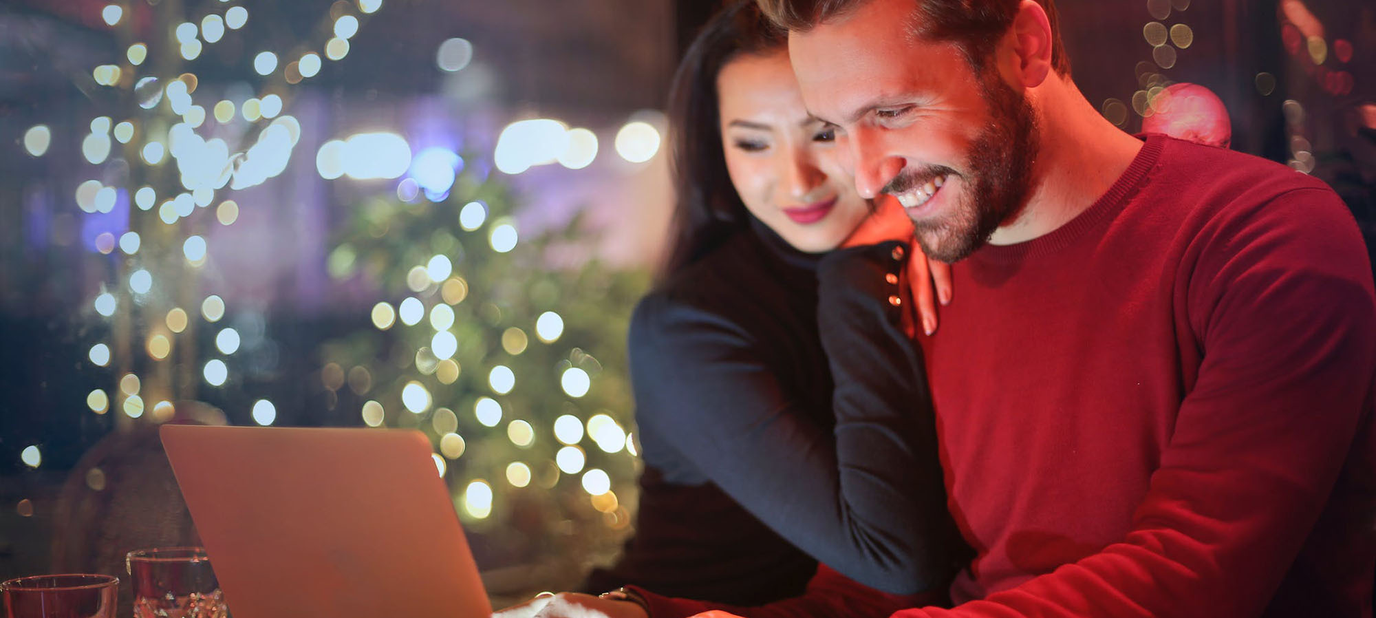 Ein Pärchen sitzt gemeinsam vor dem Laptop. Im Hintergrund sieht man weihnachtliche Dekoration, Lichterketteun, Tannenbäume.