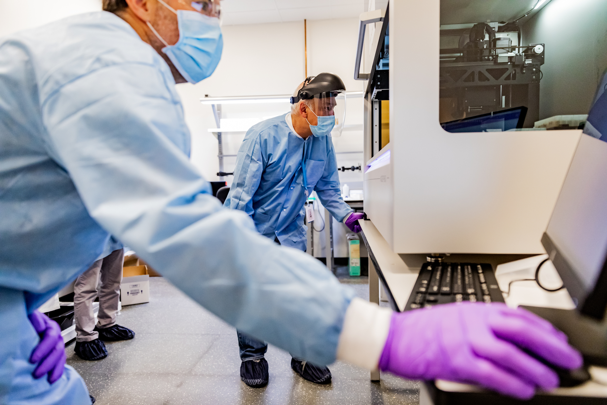 Two men working in a laboratory setting. They're wearing protective gear, including a face shield, mask, gloves, and protective clothing.