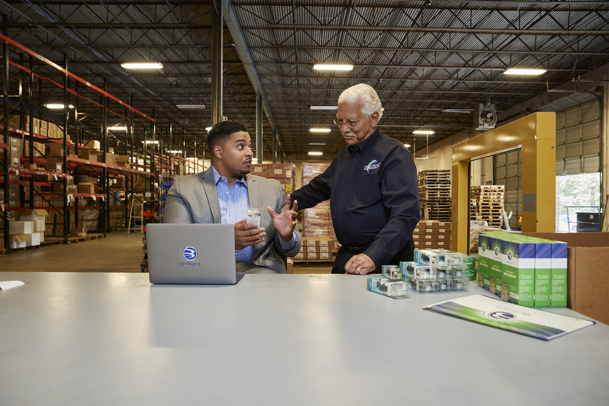 Two men speak to one another at a tabletop surface in a warehouse facility