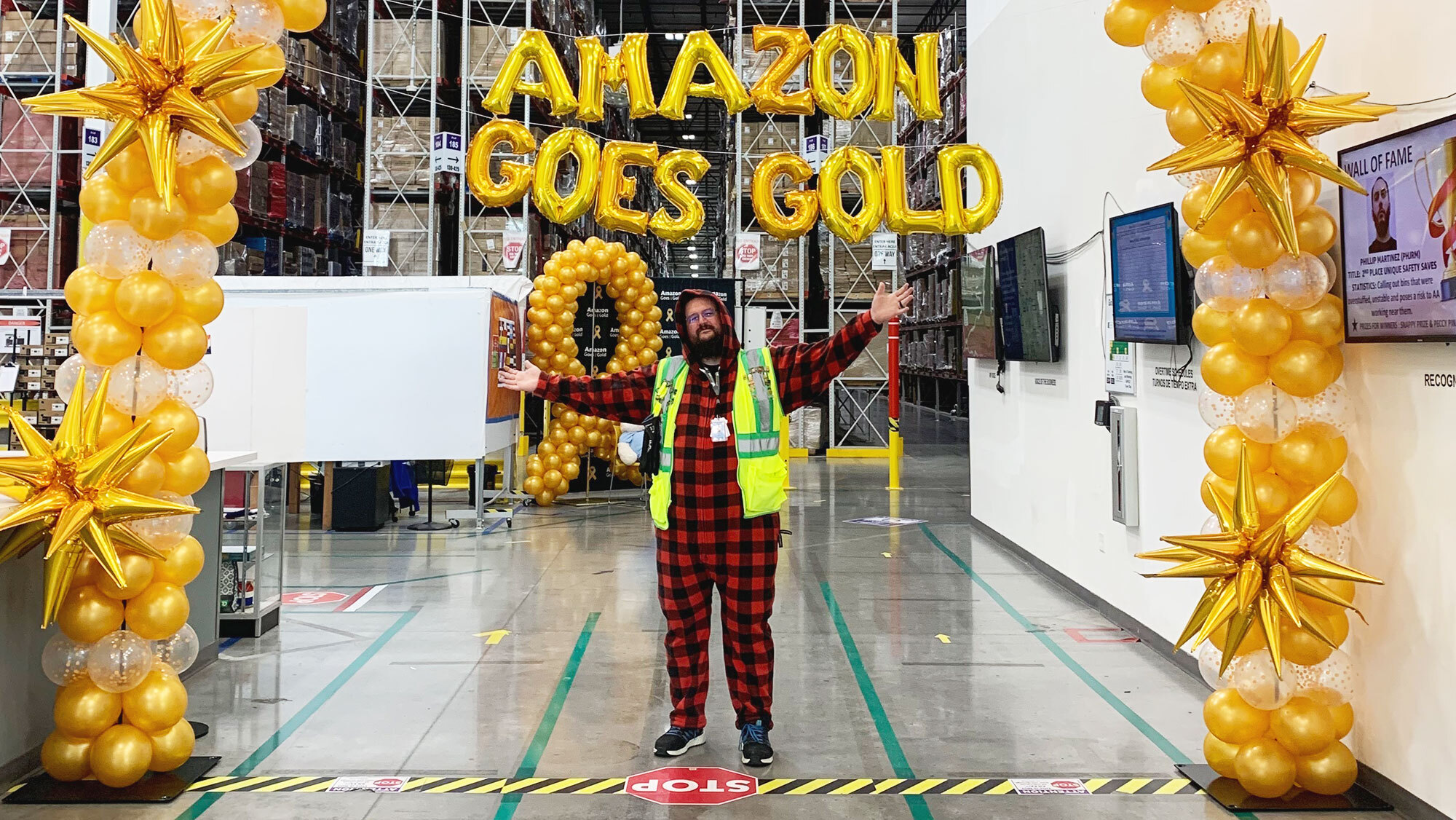 An Amazon employee wears pajamas and a yellow safety vest and stands under a balloon arch and "Amazon Goes Gold" balloon sign in a fulfillment center.