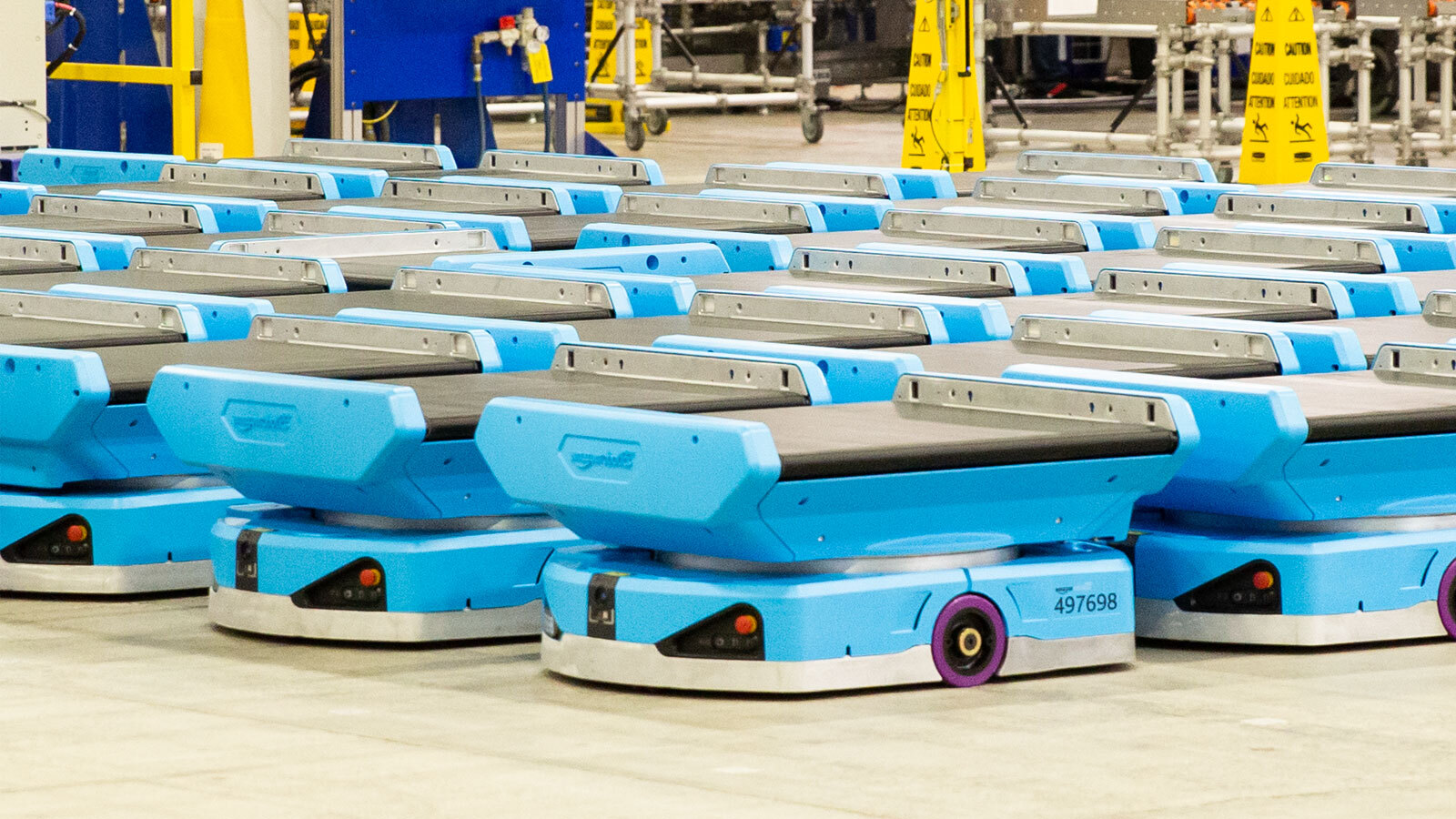 An image of multiple blue robots lined up in an Amazon fulfillment center. They are short plate-like robots with an extension plate on top that has a black conveyer belt on it. They also have cameras and sensors around the bottom plate. 