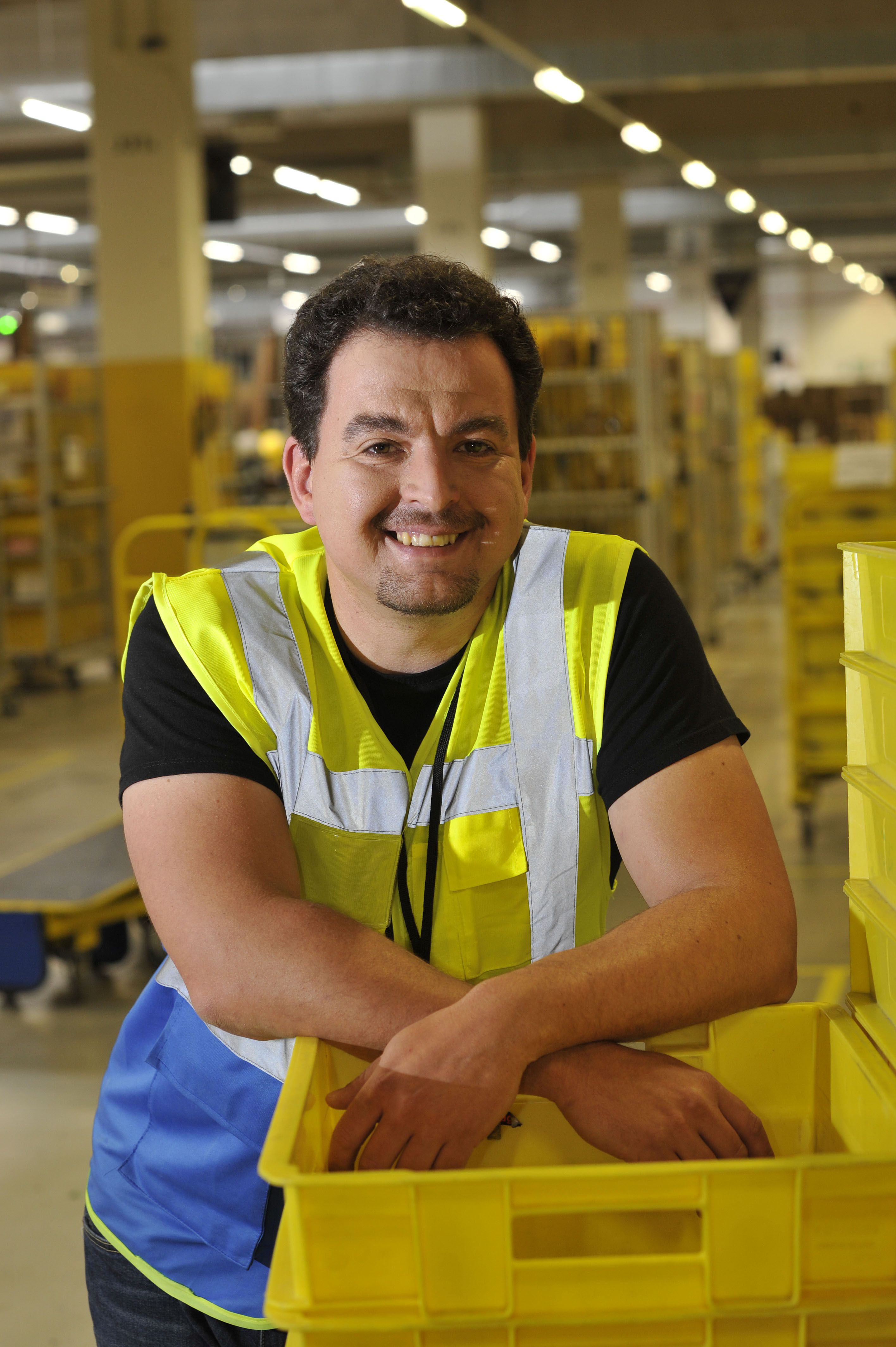 Nikolai Lisac, ein Mann mit Sicherheitsweste und braunem, gelocktem Haar, lehnt locker an einen Stapel von gelben Behältern in einem Logistikzentrum