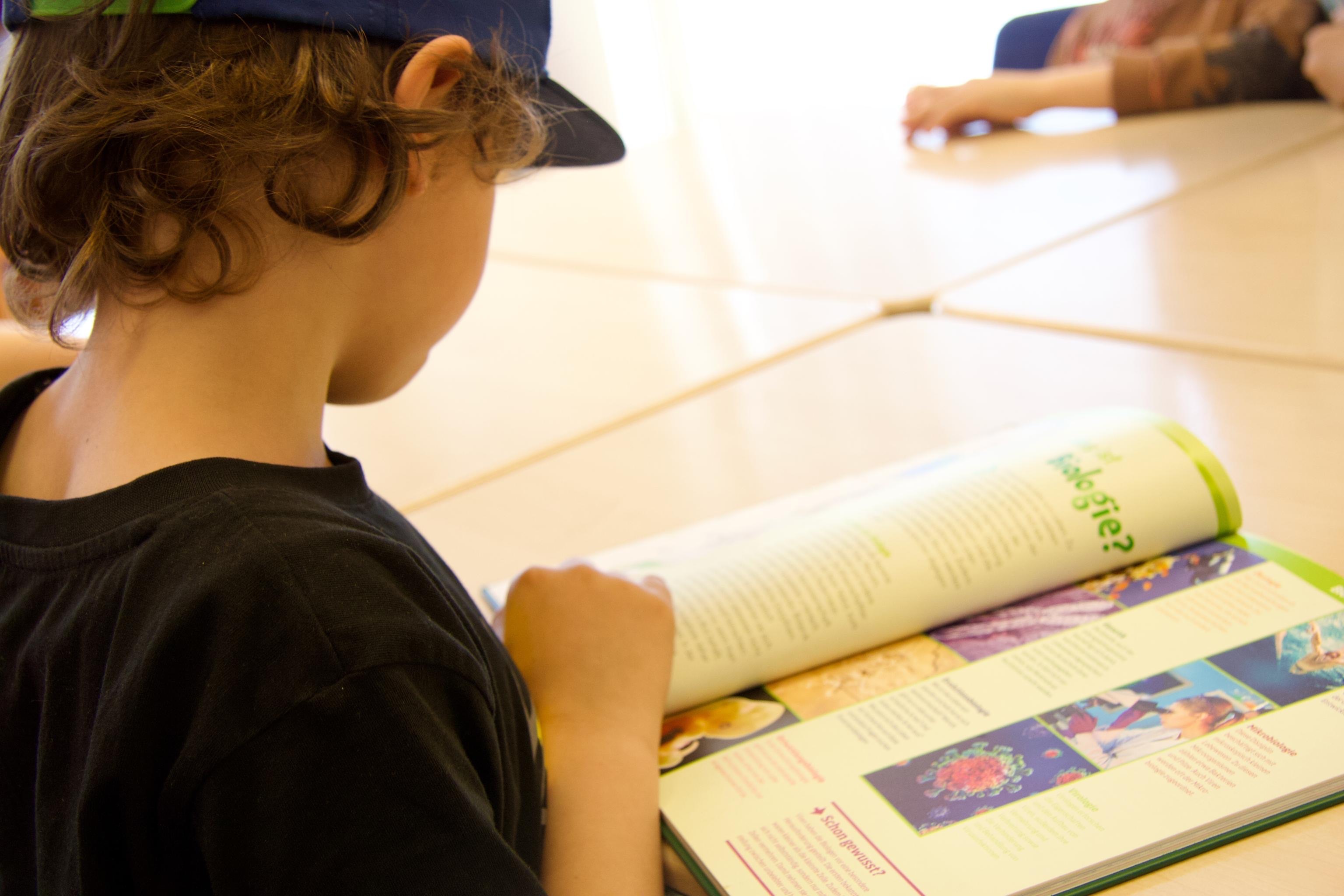 Ein Kind mit Basecap sitzt an einem Tisch und liest in einem Buch.