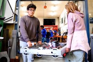 A photo of two students carrying a partially-built robot in a high school robotics lab.