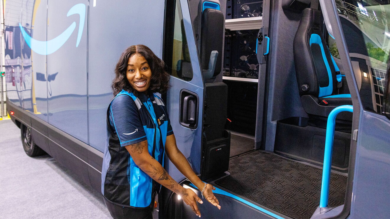 A photo of Brandi Monroe, a delivery driver for Kangaroo Direct, an Amazon Delivery Service Partner in the Baltimore, Maryland area. Brandi is standing at the front of the entrance door to an Amazon delivery van from Rivian, with arms open to the staircase.