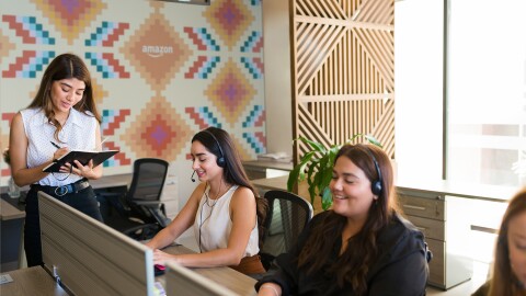 Oficina de atención al cliente de Amazon. Aparecen cuatro personas, trs sentadas en una mesa con auriculares y ordenador y una de pie apuntando en una libreta.