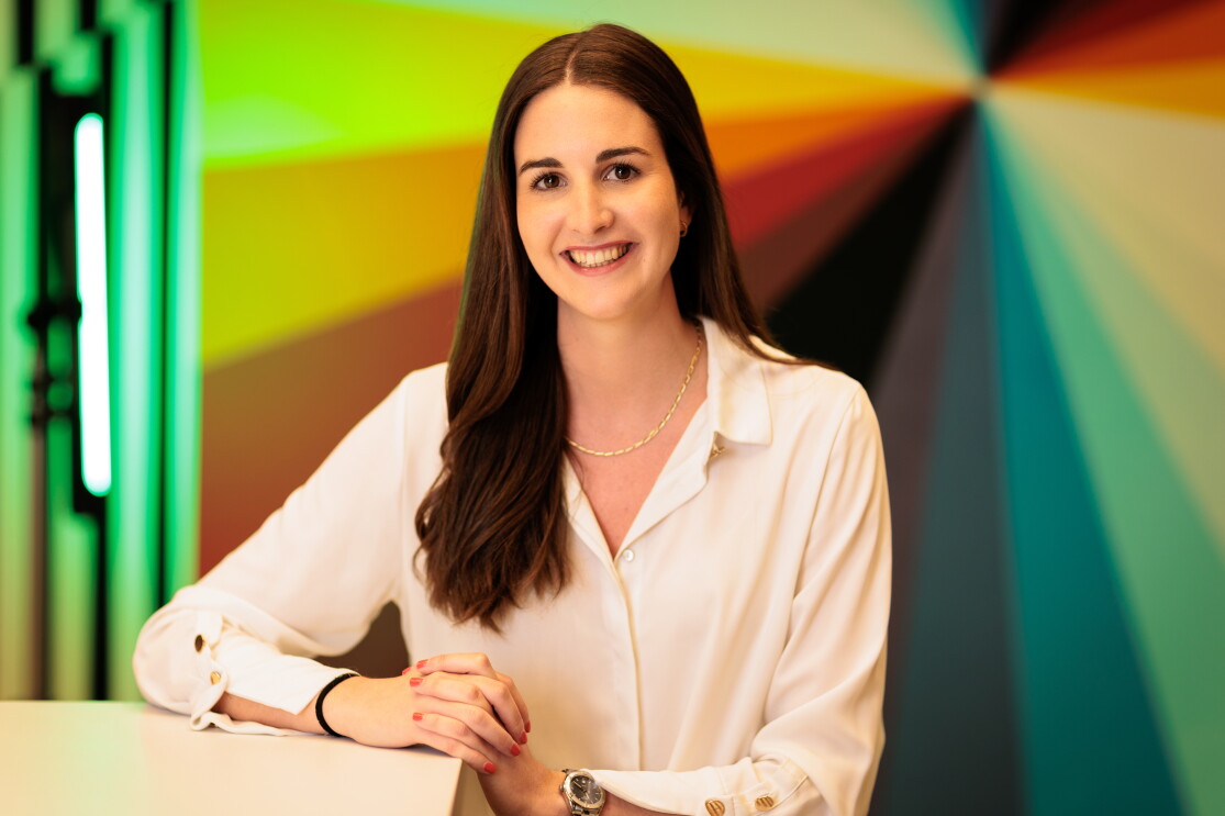 Miriam Domínguez-Adame Aguilera con el pelo liso negro hasta el pecho y una camisa de color blanco. Lleva un collar y un reloj en la mano izquierda. Un fondo de colores con un punto de fuga. Está apoyando las manos en una mesa alta. 
