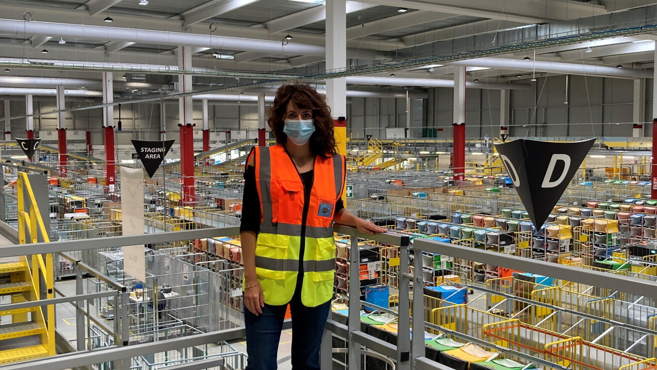 Donna in piedi su un piano rialzato in un centro operativo Amazon. Indossa una mascherina chirurgica, ha i capelli neri. 
