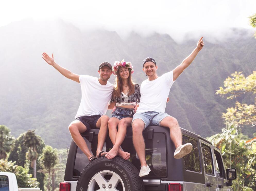 Zwei Männer und eine Frau sitzen auf einem Autodach und lachen in die Kamera. Sie trägt einen Hawaii-Haarkranz.