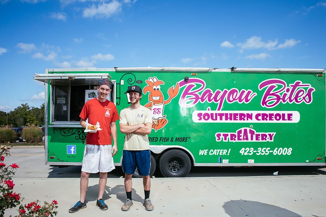 Two men stand together in front of a green food truck branded with "Bayou Bites - southern creole." The man on the left holds a basket of food, and wears a headscarf, t-shirt, basketball shorts and athletic shoes. The other man wears a baseball cap, t-shirt, basketball shorts and athletic shoes.