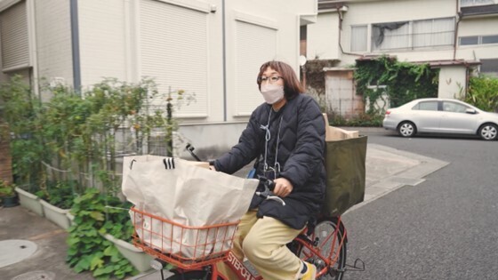 屋外、オレンジ色の自転車をこぐ女性。黒いダウンを着用。自転車の前カゴと荷台の両方に荷物が積まれている。