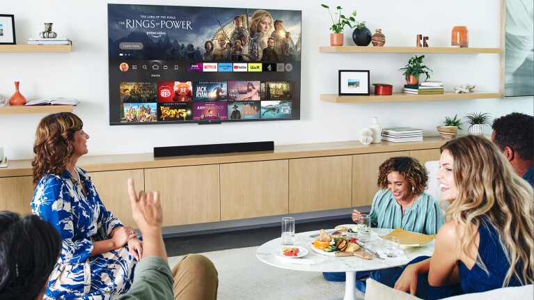 A group of people gathered in a living room with a Fire TV mounted on the wall and food on the table.