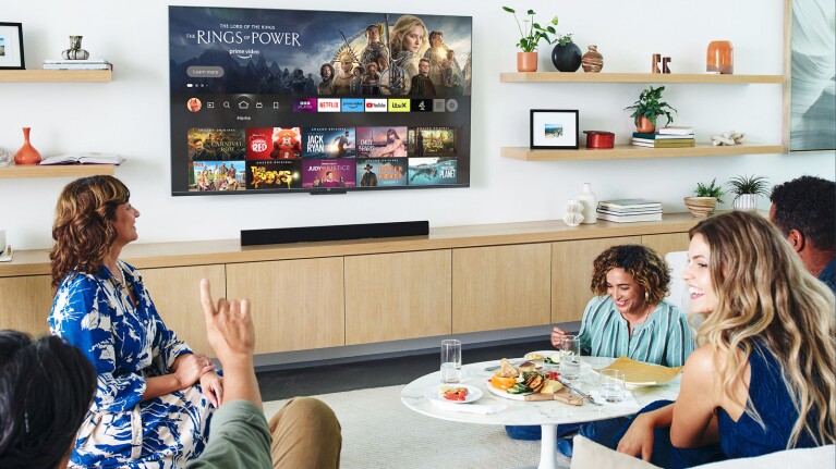 A group of people gathered in a living room with a Fire TV mounted on the wall and food on the table. 