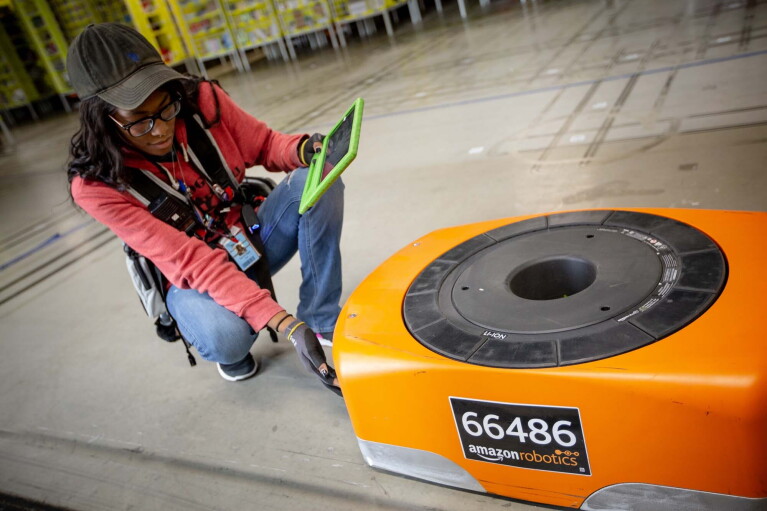 A woman kneels next to a robot, while also working on a tablet, at a Columbus, Ohio fulfillment center, CMH1