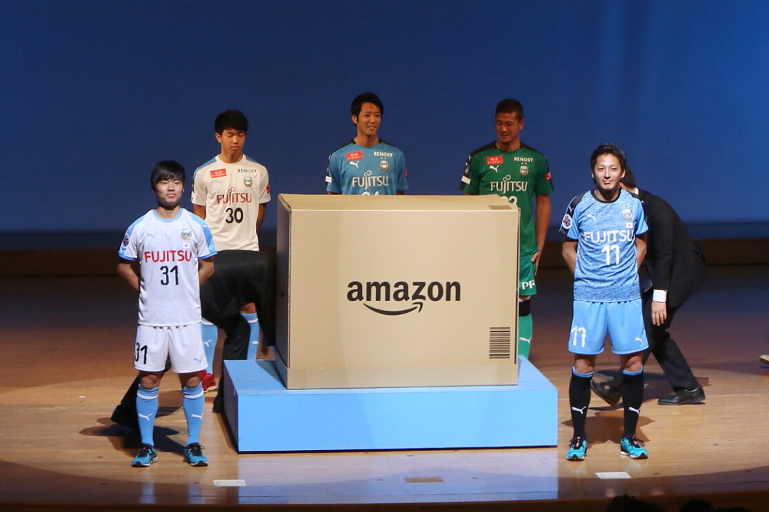 薄暗い室内、スポットライトで照らされたステージに立つ5人の男性。全員ユニフォーム姿で、Amazonロゴの入った巨大なダンボール箱を囲んでいる。