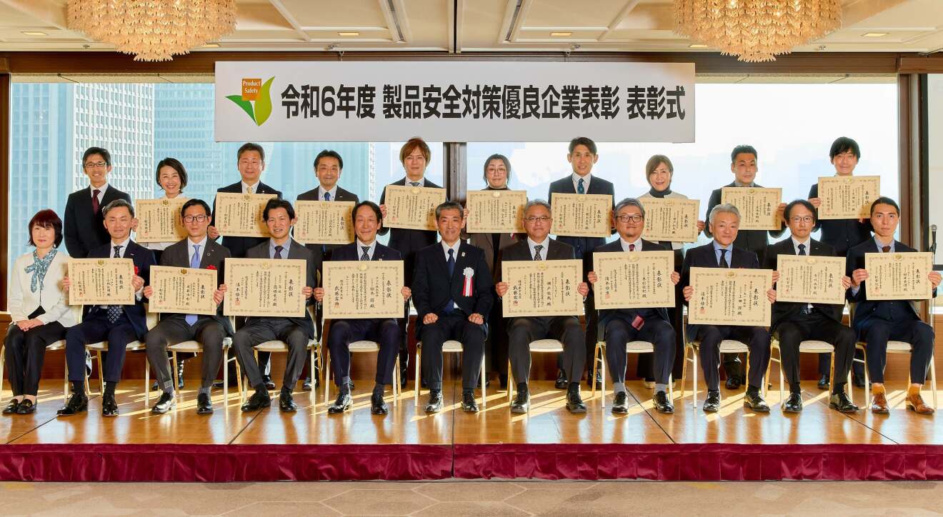 ステージにスーツを着ている男女21人が並んでいる。18人は大きな認証を持って見せられている。