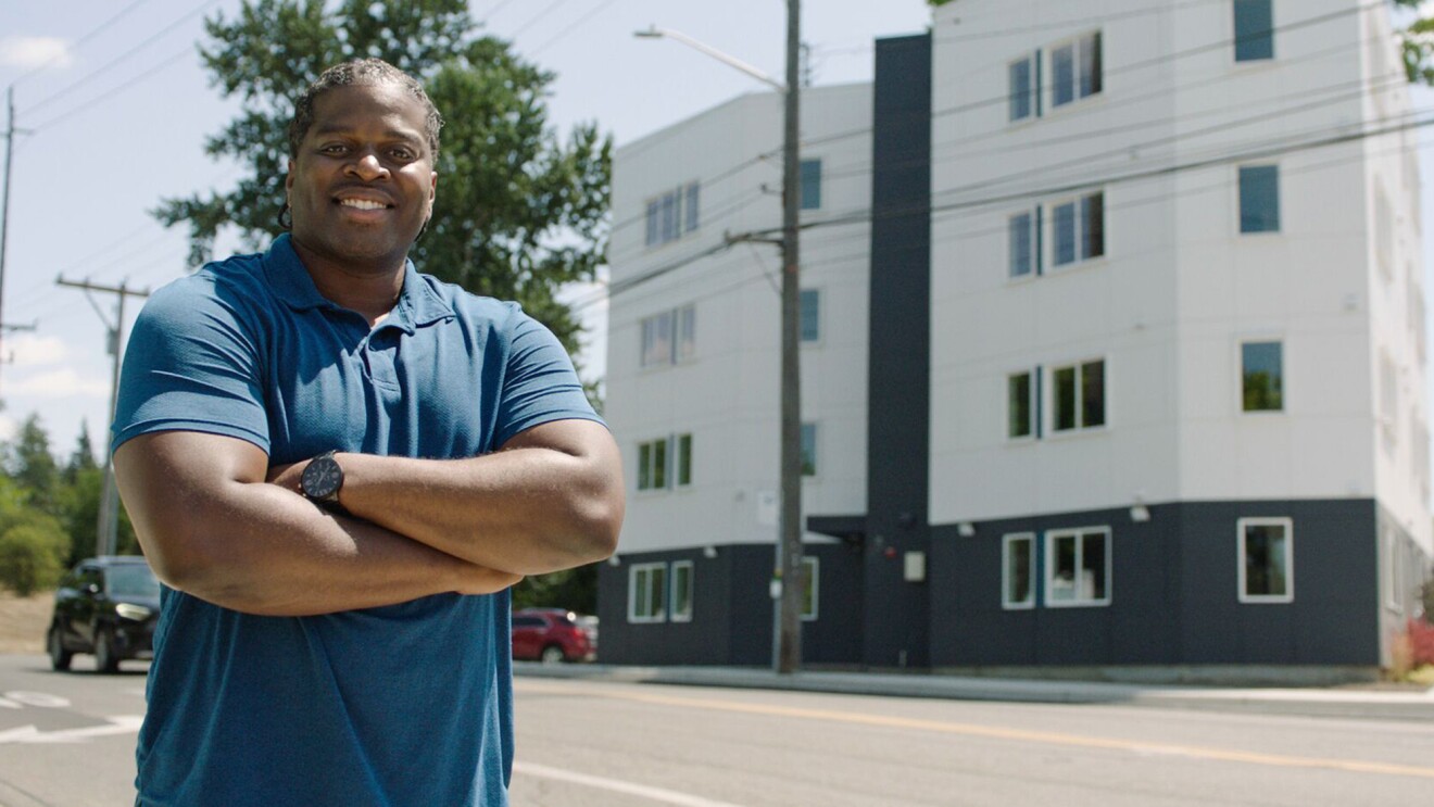 A photo of a man standing outside on a street crossing his arms. Behind him is an apartment building.