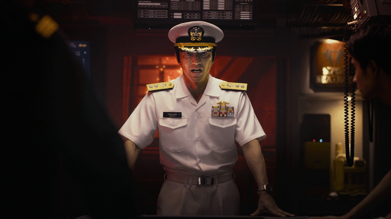 薄暗い室内で、肩に紀章のついた白い軍服を着用した男性が立っている。背後に計器類が見える。