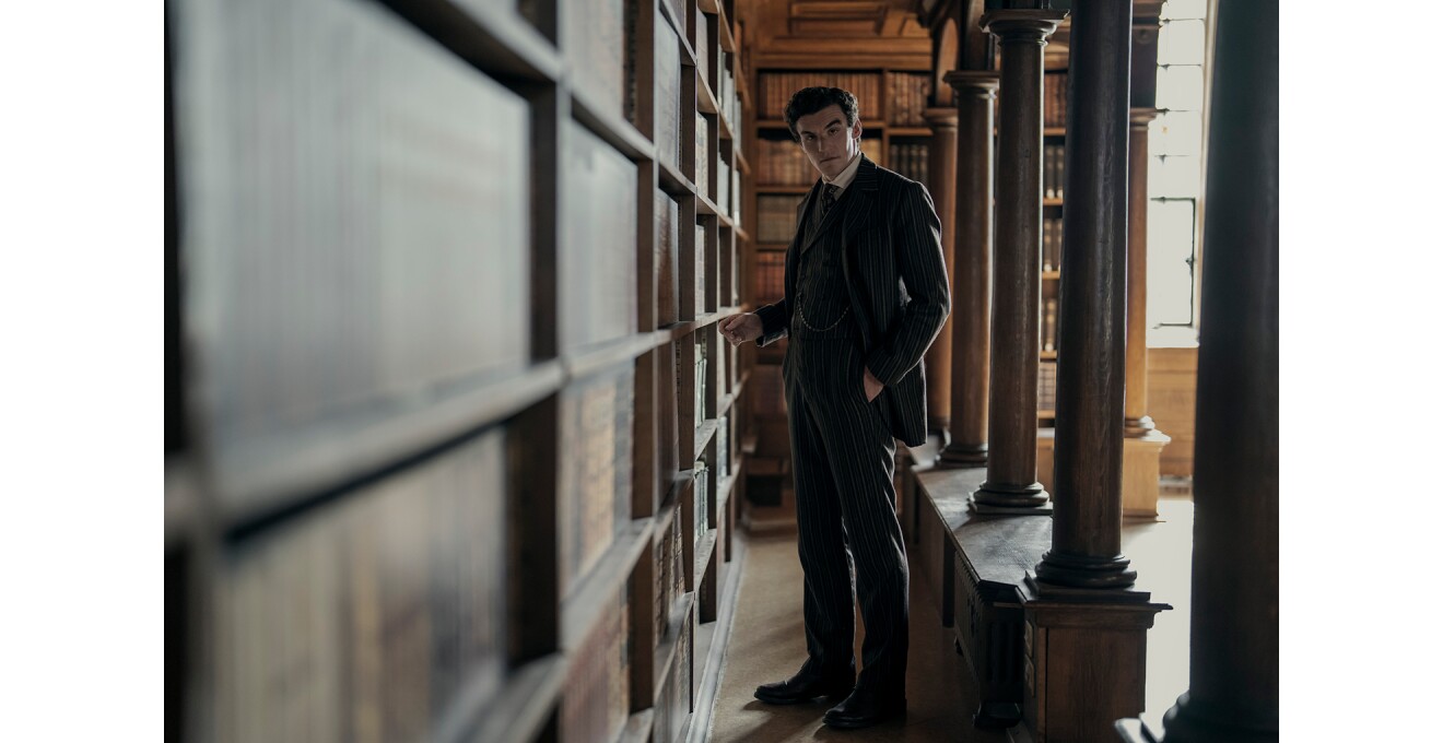 円柱と書棚が並ぶ古風な図書室に立つスーツの男性。