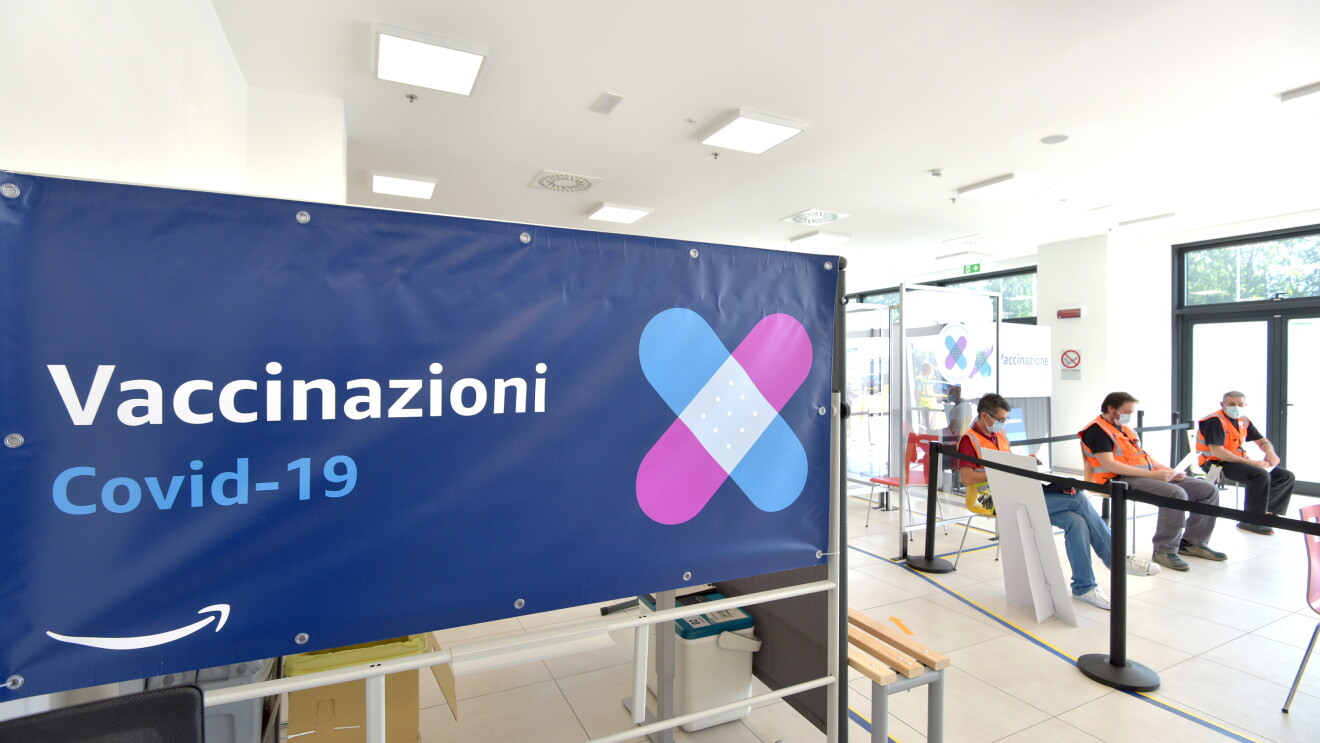 Primo piano di un cartellone blu in un sito Amazon in cui c'è scritto 'Vaccinazioni Covid-19'. Sullo sfondo, 3 dipendenti Amazon in attesa del loro turno. 