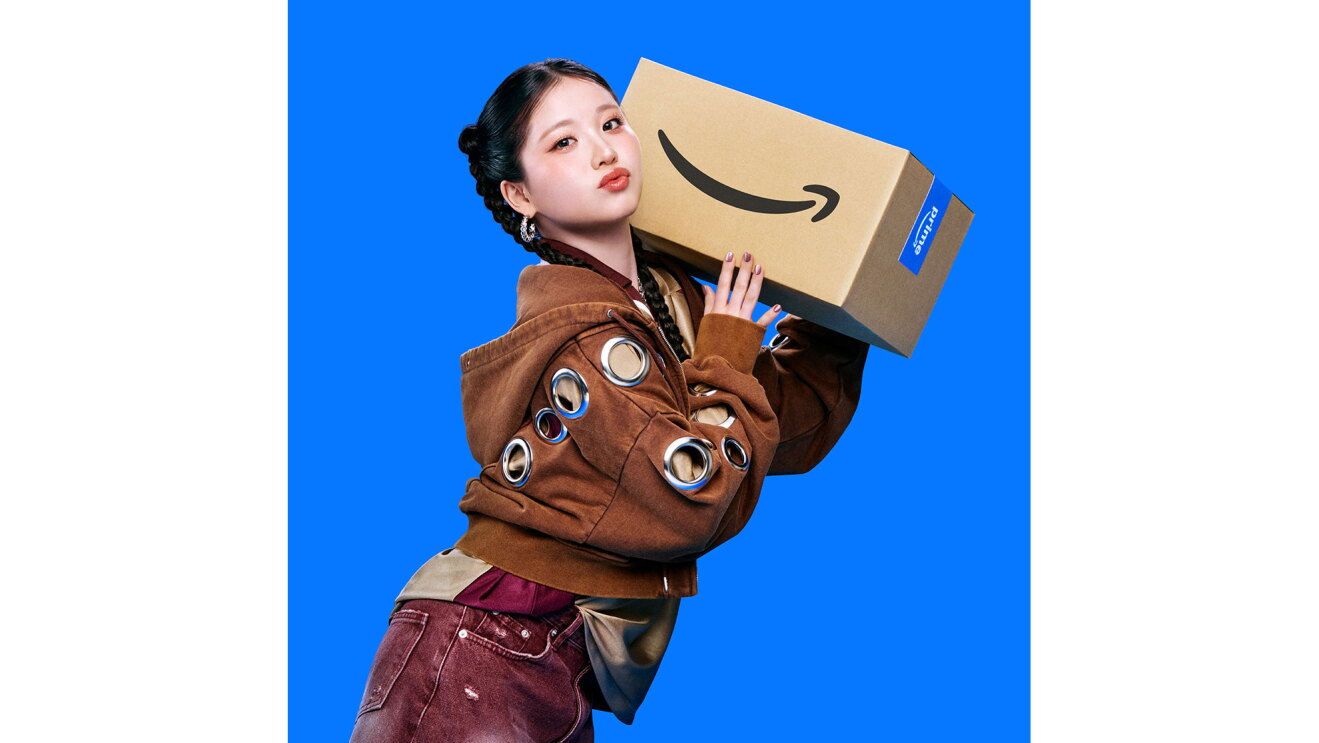 紙を三つ編みにした若い女性が、Amazonロゴ入りダンボール箱を肩に載せて両手で支えている。